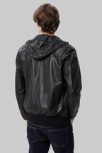 back pose Full-length studio shot of a male model wearing a modern, slim-fit black leather hooded bomber jacket over a black t-shirt and dark blue jeans against a solid white background.