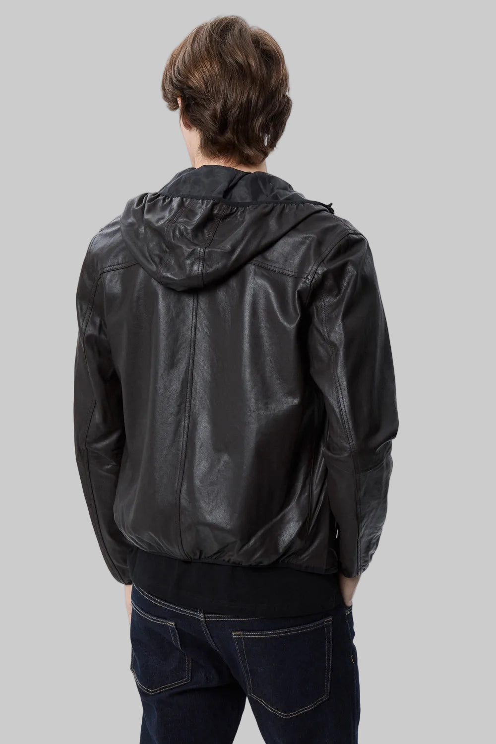 back pose Full-length studio shot of a male model wearing a modern, slim-fit black leather hooded bomber jacket over a black t-shirt and dark blue jeans against a solid white background.