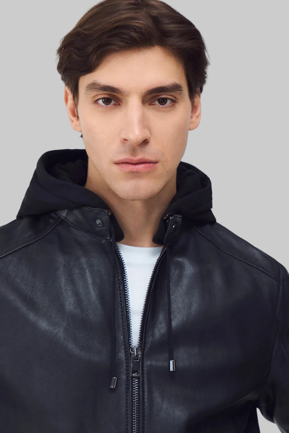 Front studio shot of a man wearing a slim-fit black leather bomber jacket with a snap-tab collar and zippered pockets, styled with dark pants against a white background.
