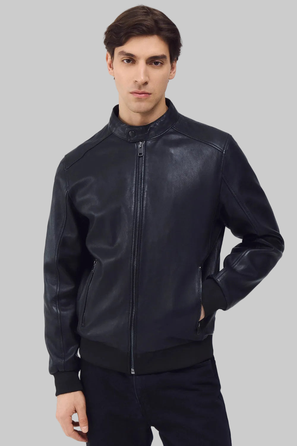 Front studio shot of a man wearing a slim-fit black leather bomber jacket with a snap-tab collar and zippered pockets, styled with dark pants against a white background.