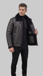 A man wearing a black leather jacket with a black shearling collar, standing in an industrial loft setting. He is holding the jacket open to show the interior lining and pocket details, which feature the brand name 'LTX'.