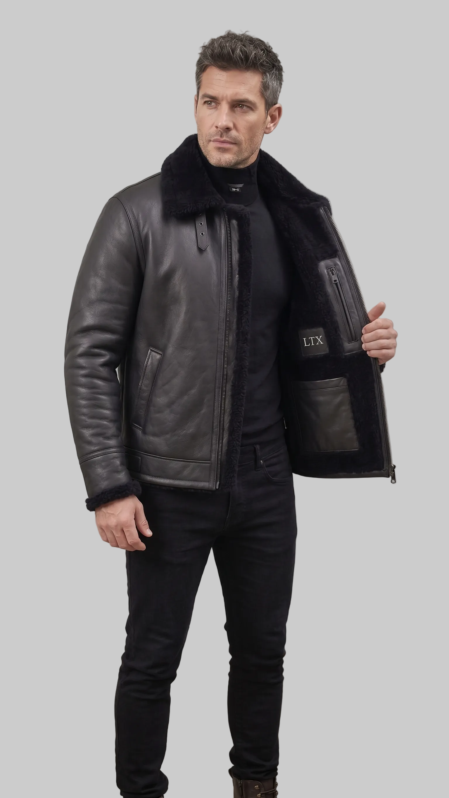 A man wearing a black leather jacket with a black shearling collar, standing in an industrial loft setting. He is holding the jacket open to show the interior lining and pocket details, which feature the brand name 'LTX'.