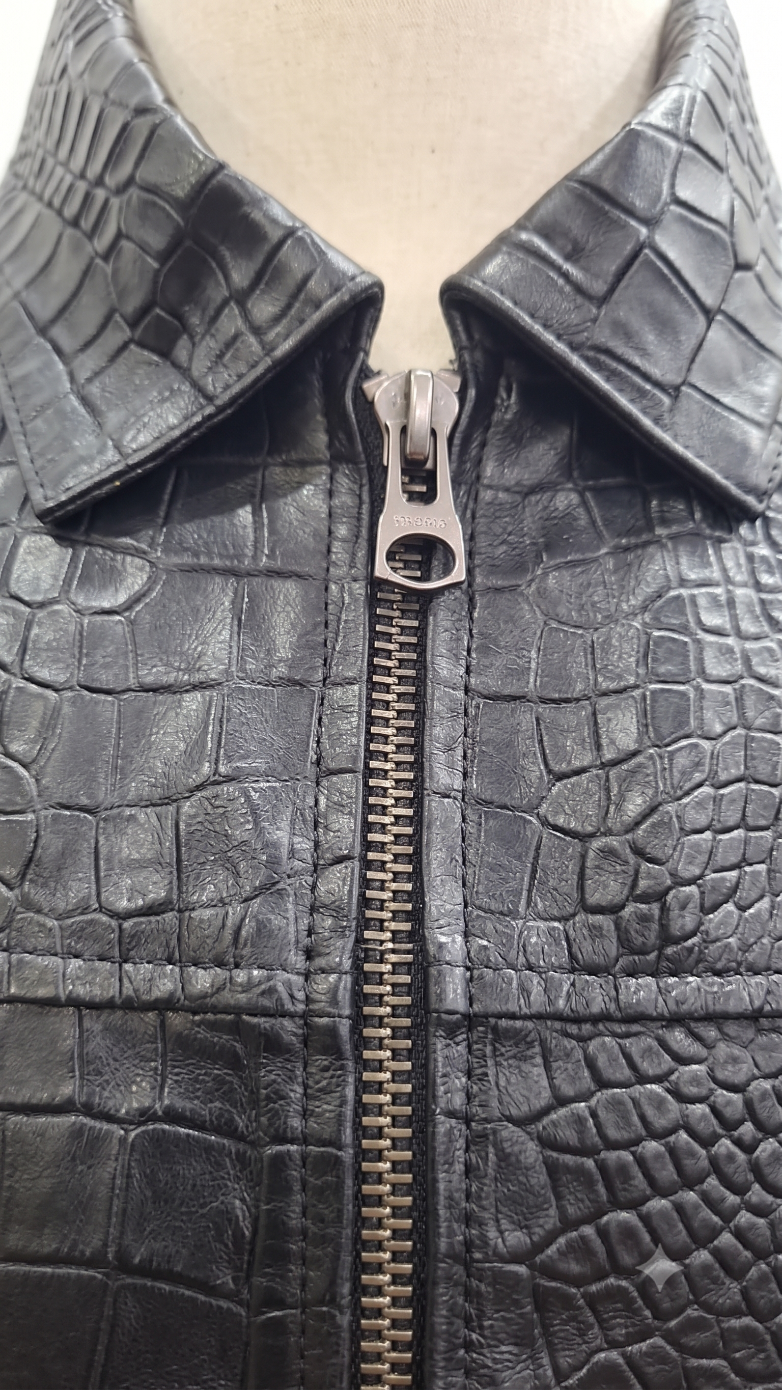 Close-up of a black crocodile-embossed leather jacket with a silver metal zipper, displayed on a beige mannequin torso.