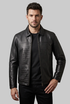 A man wearing a black textured leather jacket with a crocodile-embossed pattern over a black t-shirt, standing against a textured gray studio background.