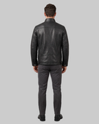 A man viewed from behind wearing a black leather jacket, dark gray slim-fit trousers, and black shoes, standing against a light gray studio background.