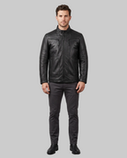 A man modeling a black leather jacket with zippered chest pockets, paired with dark gray trousers and black shoes, standing against a solid gray studio background.