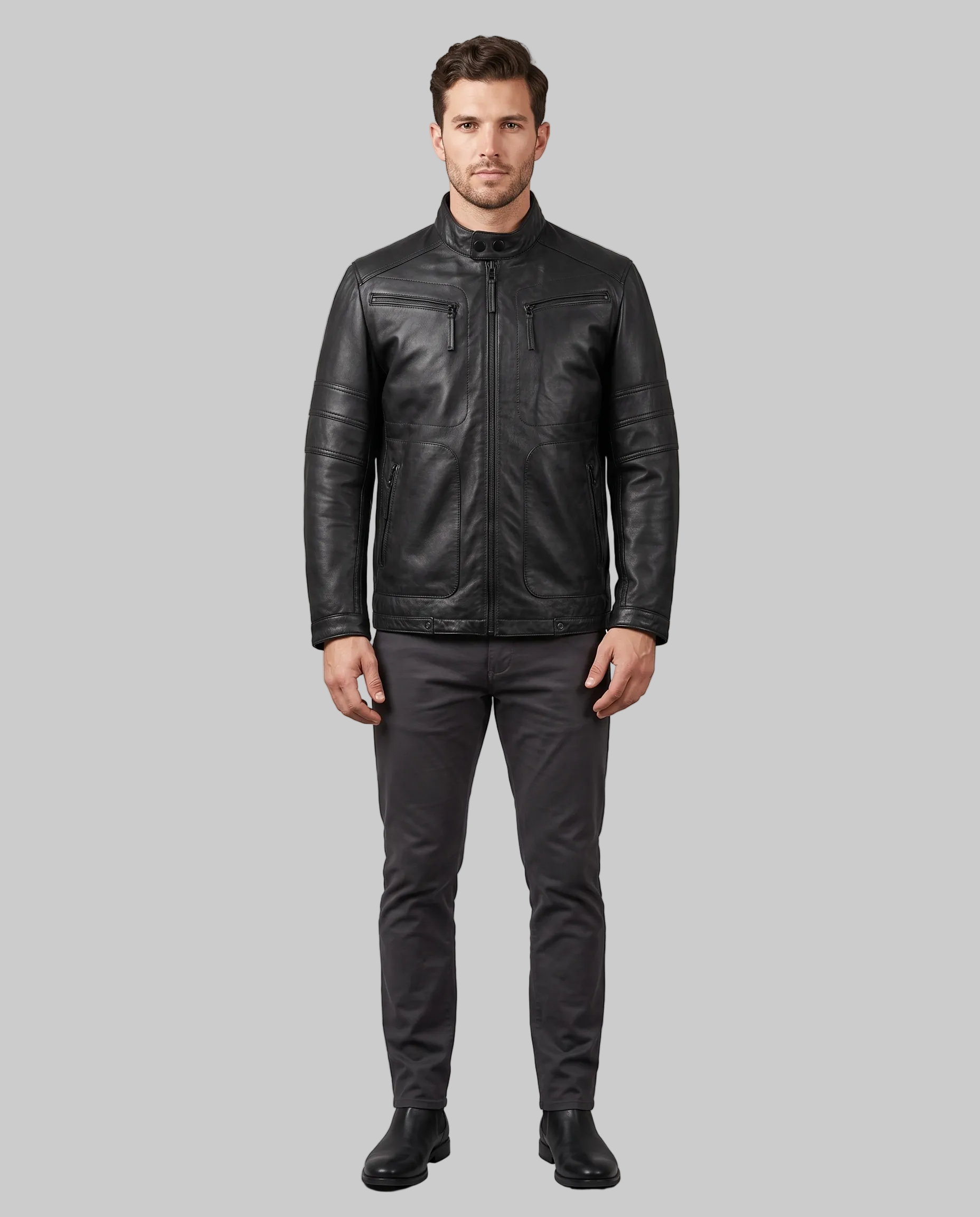 A man modeling a black leather jacket with zippered chest pockets, paired with dark gray trousers and black shoes, standing against a solid gray studio background.