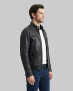 A man wearing a black leather motorcycle jacket over a white t-shirt and blue jeans, shown in profile against a light gray studio background.