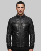 A man wearing a black leather motorcycle jacket with a front zipper, zippered chest pockets, and ribbed cuffs, photographed against a plain light gray studio background.