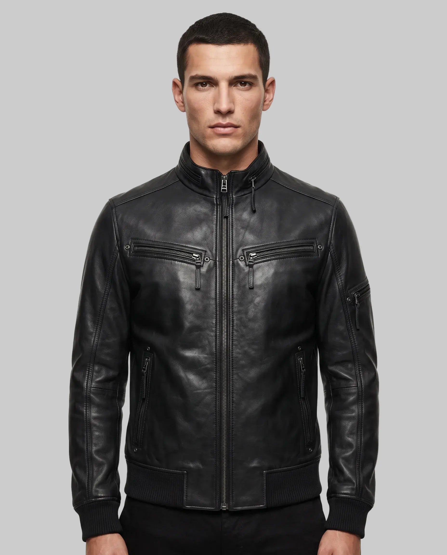 A man wearing a black leather motorcycle jacket with a front zipper, zippered chest pockets, and ribbed cuffs, photographed against a plain light gray studio background.