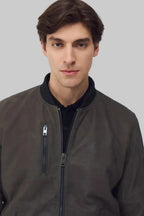 A man wearing a dark brown suede bomber jacket with a black ribbed collar, shown from the chest up against a solid gray background.