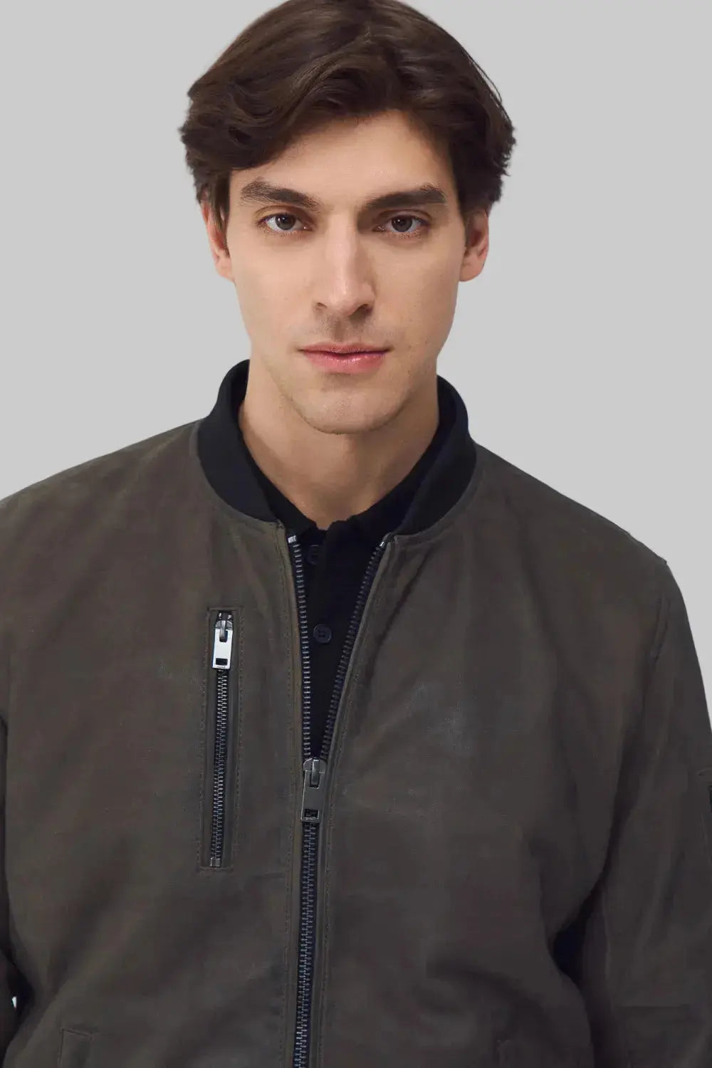 A man wearing a dark brown suede bomber jacket with a black ribbed collar, shown from the chest up against a solid gray background.