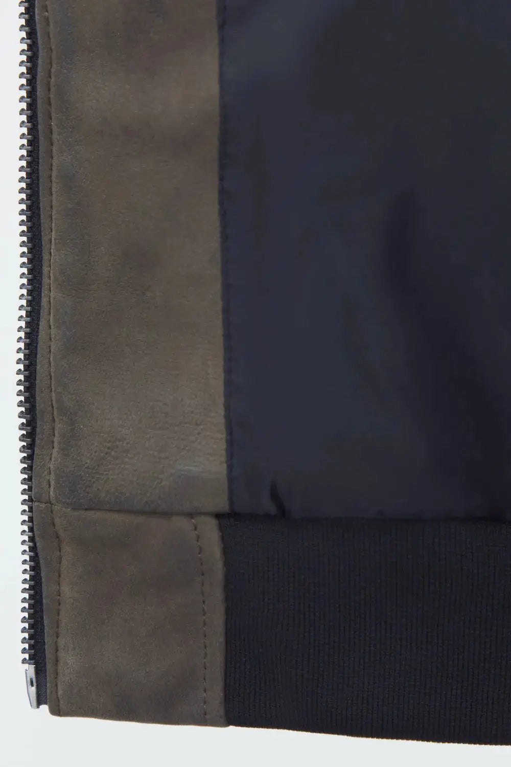 Close-up detail of a patchwork jacket showing a dark navy nylon body, a taupe-brown suede panel, and a black ribbed knit cuff with a silver zipper.