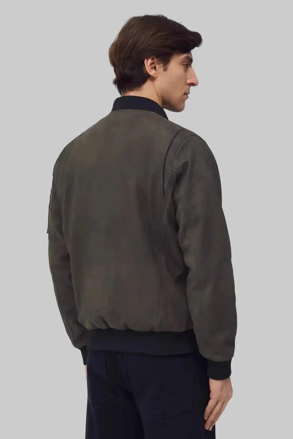A man shown from the back wearing a dark olive-brown suede bomber jacket with black ribbed trim, paired with dark trousers, against a solid gray background.