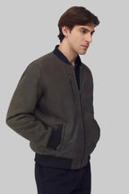 A man wearing a dark olive green zip-up bomber jacket with black ribbed trim, shown from the waist up against a solid gray background.