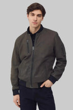 A man wearing a dark olive-brown bomber jacket with black ribbed trim, standing against a solid gray studio background.