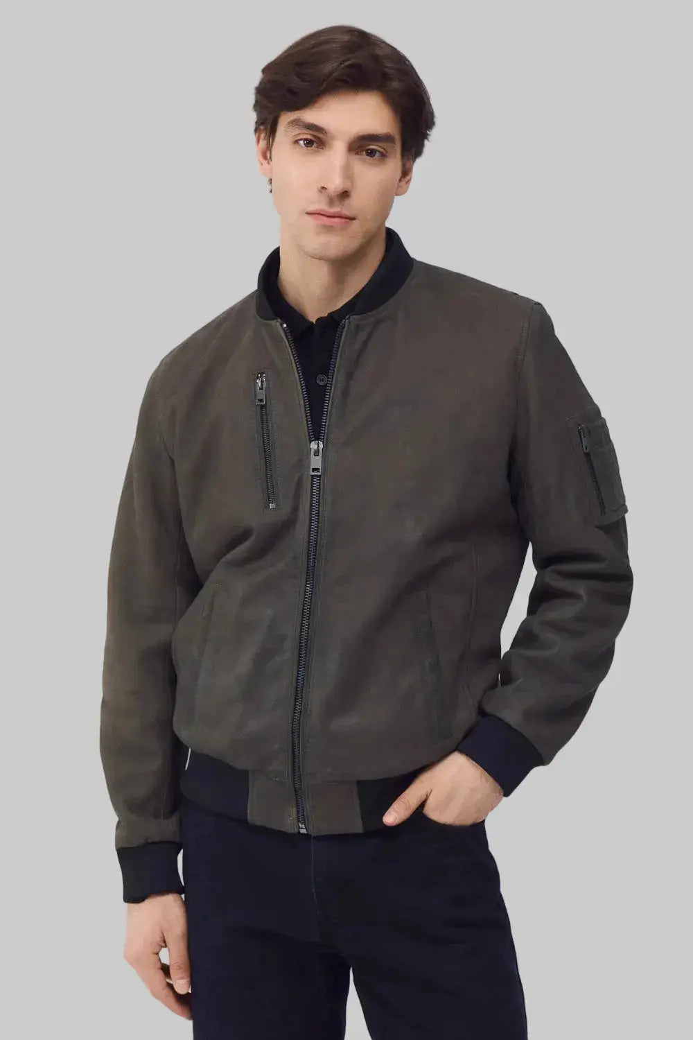 A man wearing a dark olive-brown bomber jacket with black ribbed trim, standing against a solid gray studio background.
