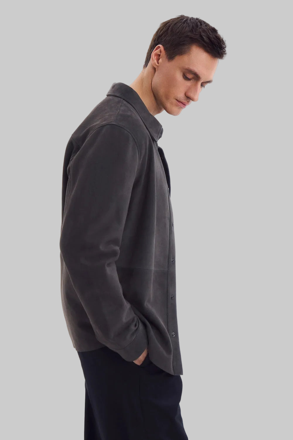 Front studio portrait of a man wearing a minimalist, charcoal gray suede shirt jacket snapped completely closed, featuring a clean concealed placket and point collar against a solid white background.